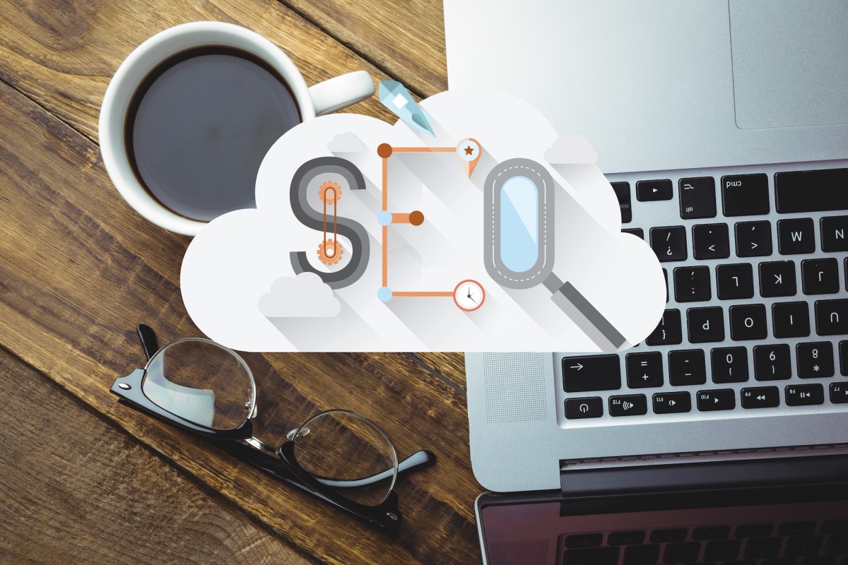 Overhead View Of Laptop, Coffee, And Glasses On A Wooden Desk, With The Text “Seo” Written Creatively In A Cloud Shape