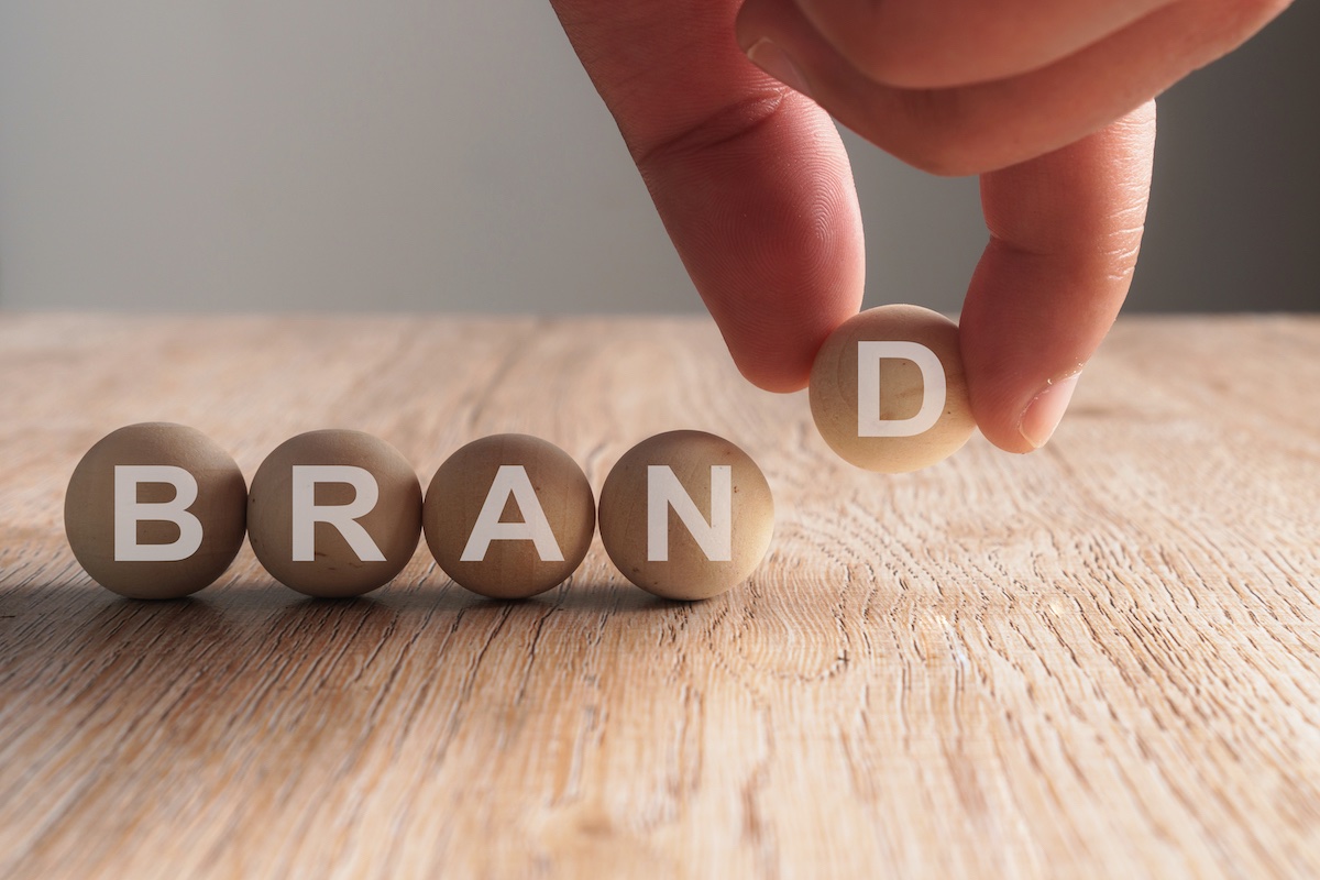 5 Wooden Balls On A Desk That Spell Out The Word “Brand”. A Hand Is Putting The Letter “D” In Place