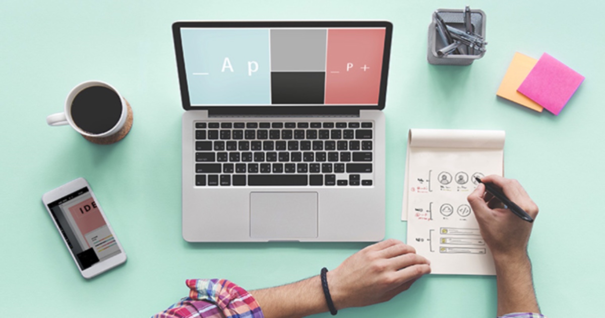 Overhead View Of Laptop On Green Desk With Phone, Coffee, And Stationery. A Man Is Working On Web Design On A Notepad.