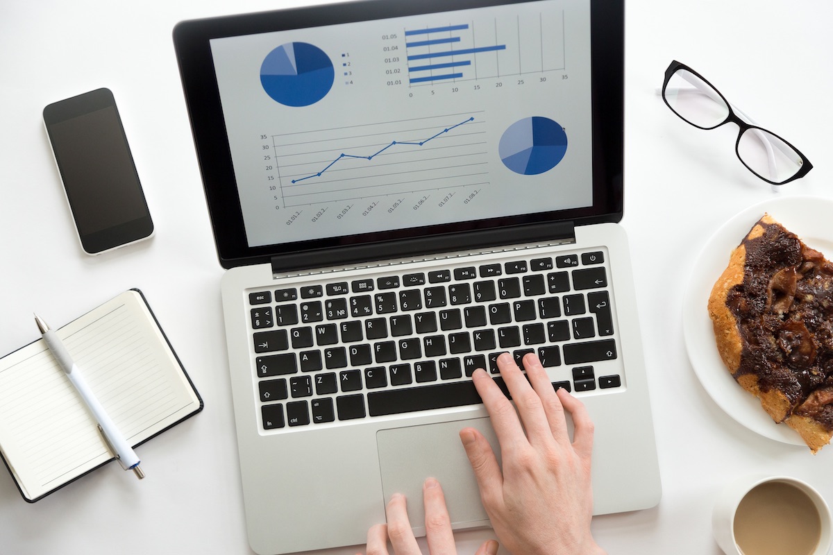 Hands On Laptop With Website Metrics Data On The Screen. Surrounding The Laptop Are A Journal And Pen, A Phone, Glasses, And A Pastry With Coffee