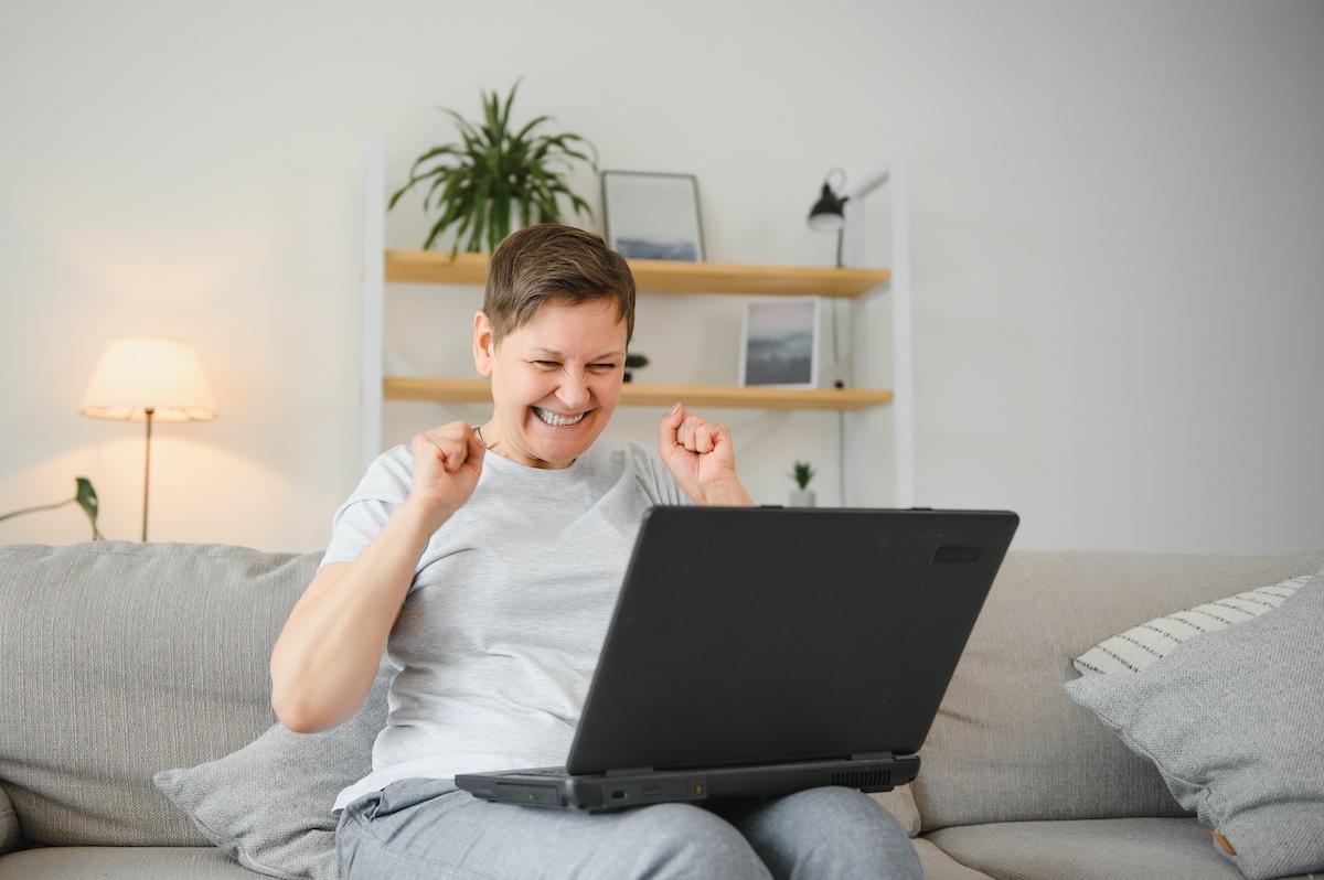 Happy Elderly Lady Sitting On Her Couch With Laptop On Her Lap. Excited About Article About How To Get Your Website Noticed