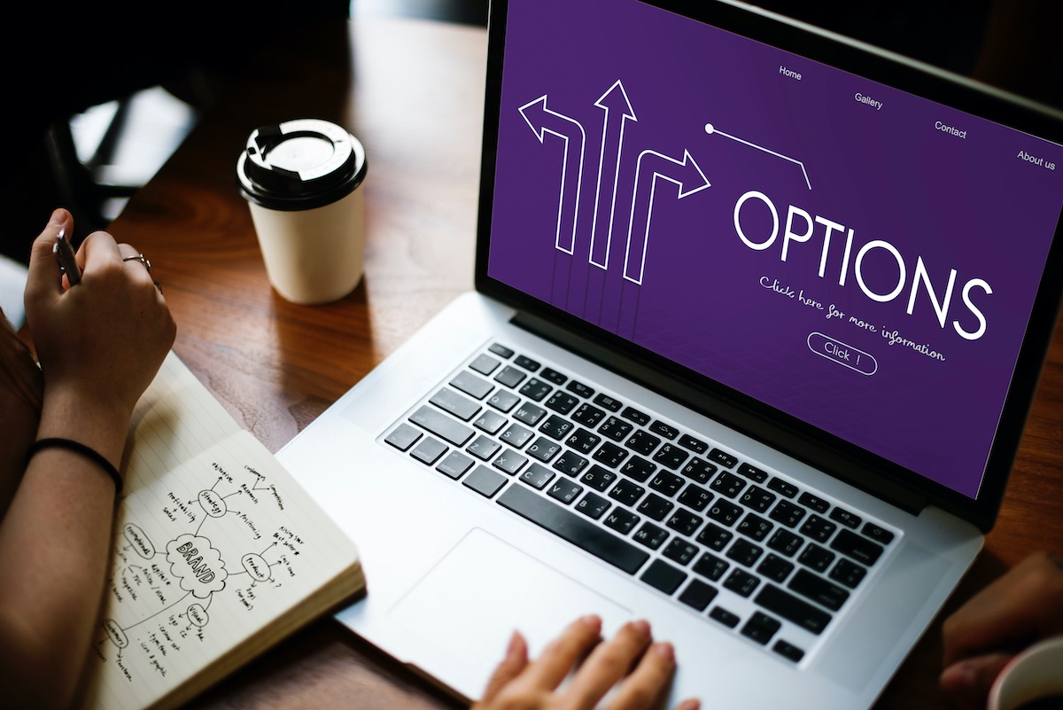 Content Writing 1 Laptop On Desk With A Purple Screen With The Text “Options” On It. There Is A Coffee Beside The Laptop And A Hand Content Writing In A Notebook