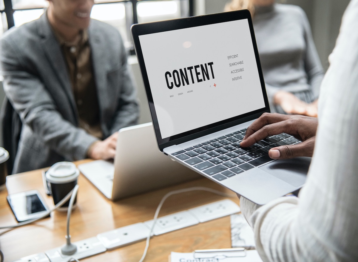 Man Holding A Laptop With “Content” Written On The Screen, Among A Busy Office Desk With Other People Working In The Background