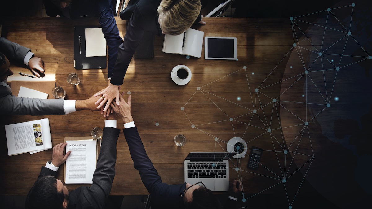 Overhead View Of A Wooden Desk With Laptops, Coffee, Water Glasses, And Clipboards On It. 5 Business Men Are Putting Their Hands Together In The Middle Of The Desk To Show Teamwork