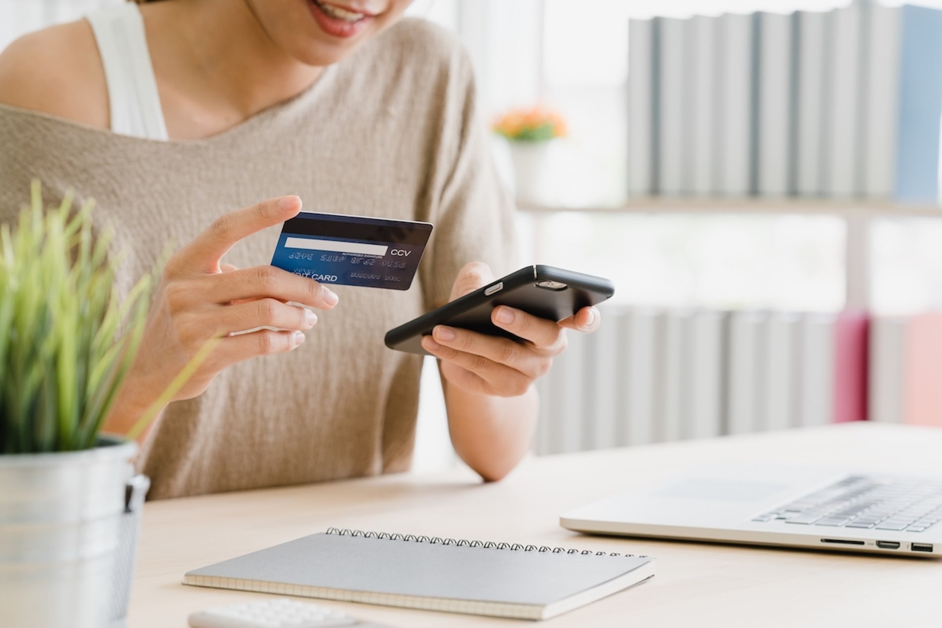 Woman in front of her laptop using credit card to purchase something on her mobile phone 